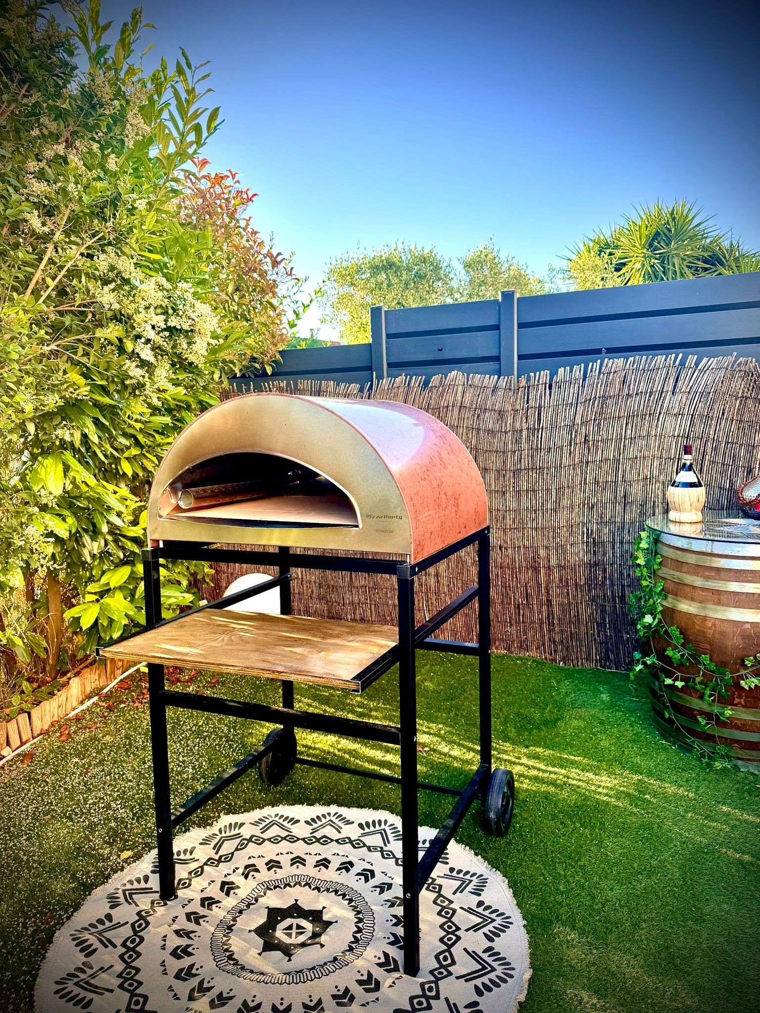 Large antique copper pizza party oven on a wooden shelf cart, in a garden with a green lawn