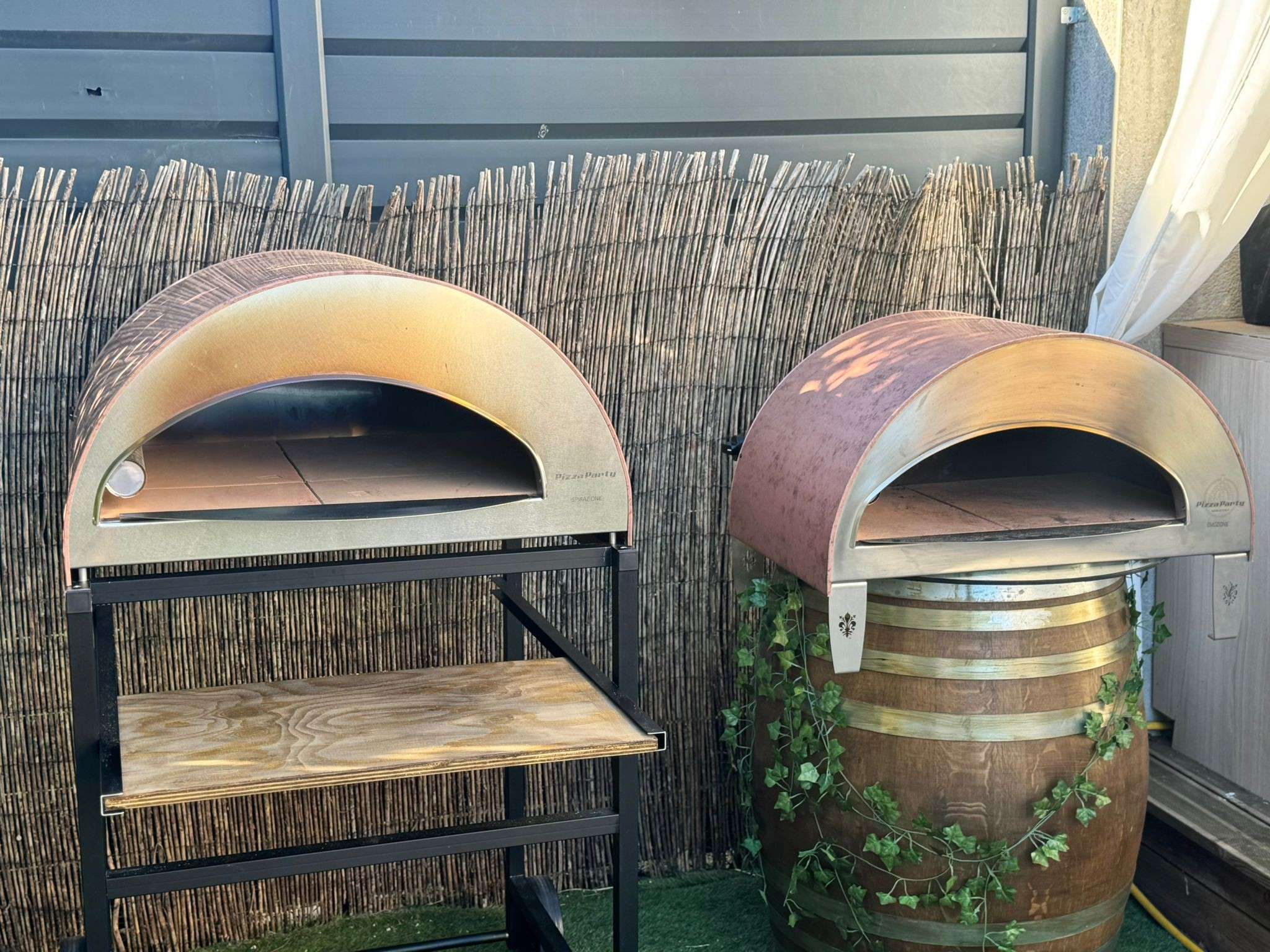 Right side view of a large antique copper Pizza Party-inspired oven on a wooden stand with a shelf, in a garden