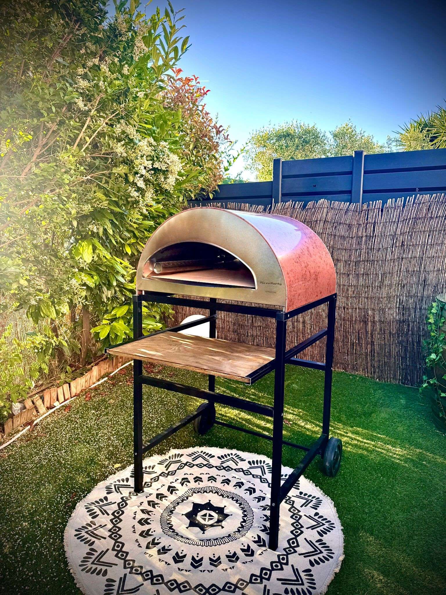 Front view of a large antique copper Pizza Party-inspired oven on a wheeled cart, set in a garden with a decorative barrel in the background