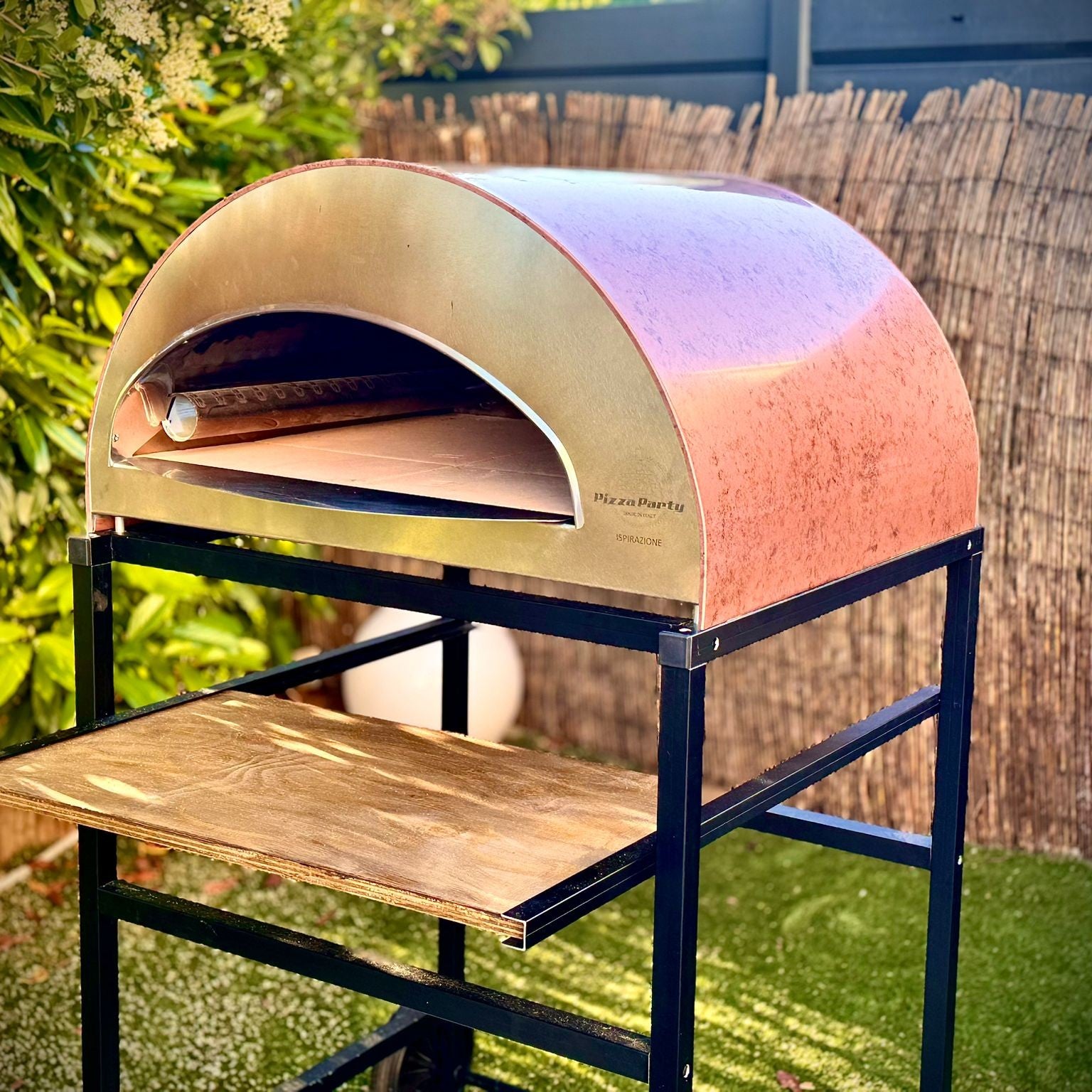 Forno Pizza Party Ispirazione Grande rame antico con piano cottura 60x70cm, vista laterale in giardino con testo "NEW 70x60cm"