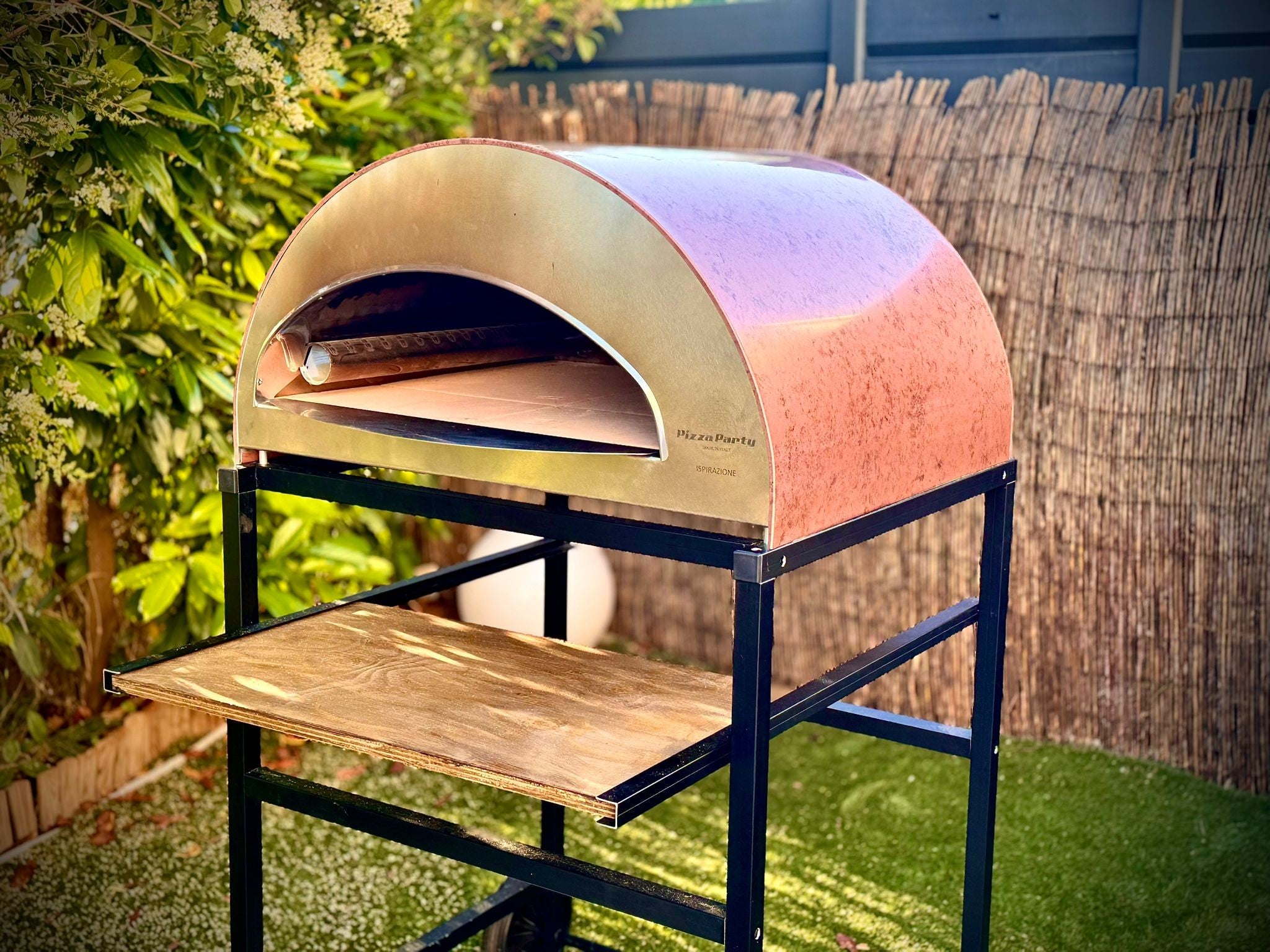 Forno Pizza Party Ispirazione Grande rame antico con piano cottura 60x70cm, vista laterale in giardino con testo "NEW 70x60cm"