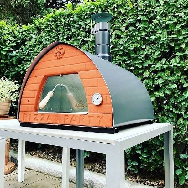 Houtgestookte Pizza Party-forno 70x70 in geweerstaalkleur op een witte tafel in de tuin met heg, vooraanzicht met thermometer