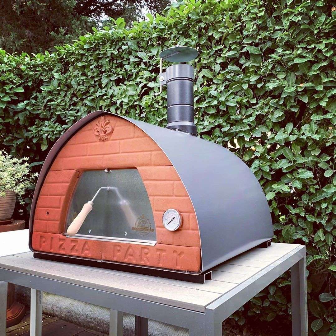 Houtgestookte Pizza Party 70x70 'Pietra' pizzaoven van boven gezien in de tuin op een witte tafel, met Vesuvius-pizzascheppen op een rotan eettafel geplaatst