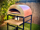 Forno Pizza Party Ispirazione Grande rame antico con piano cottura 60x70cm, vista laterale in giardino con testo "NEW 70x60cm"