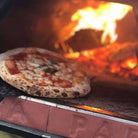 Horno de leña Pizza Party 70x70 "Piedra" sobre mesa blanca en el jardín, vista frontal con chimenea posterior